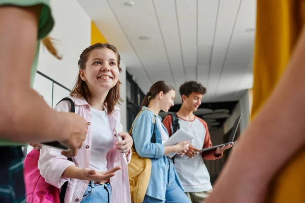 De vuelta a la escuela, chica adolescente feliz hablando con compañeros de clase en el pasillo, estudiantes borrosos utilizando dispositivos - foto de stock