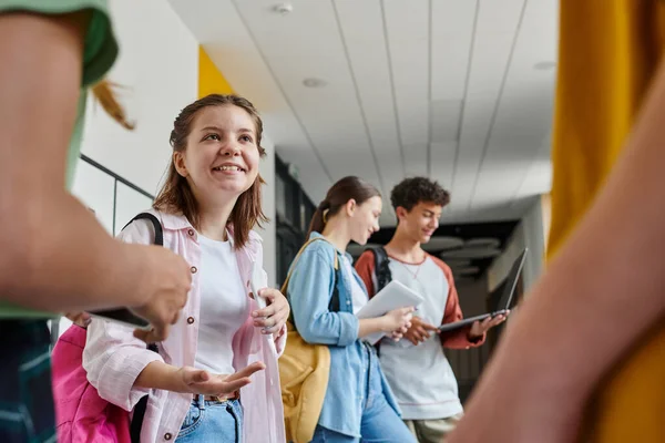 De vuelta a la escuela, chica adolescente feliz hablando con compañeros de clase en el pasillo, estudiantes borrosos utilizando dispositivos - foto de stock