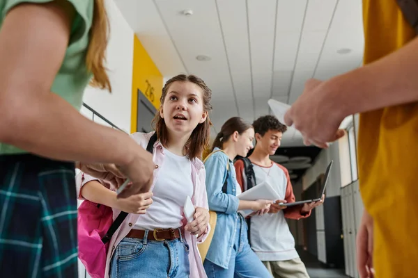 Écolière surprise regardant ses camarades de classe dans le couloir de l'école, les adolescents avec des appareils — Photo de stock