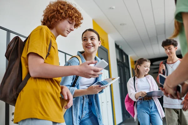 Назад в школу, одноклассники, использующие гаджеты в школьном коридоре, рыжий мальчик, использующий смартфон рядом с девушкой — стоковое фото