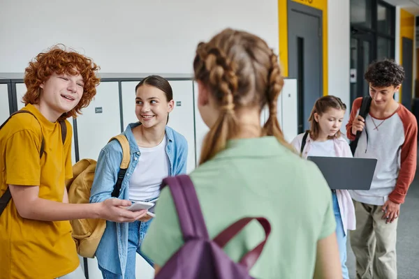Happy redhead boy holding smartphone and looking at girl on blurred foreground, teen students — Stock Photo
