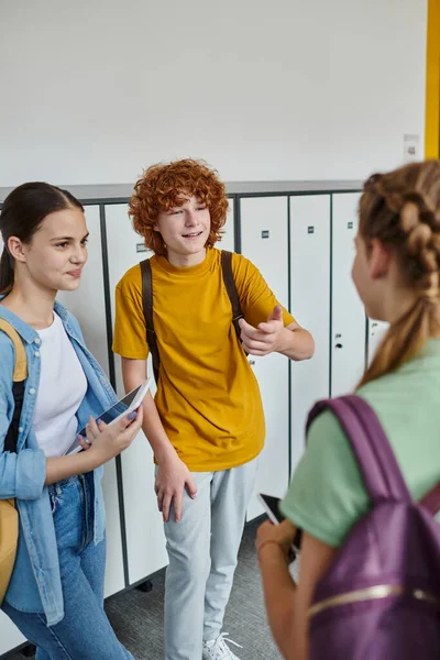 Щасливий хлопчик жестикулює і спілкується з однокласницею в шкільному коридорі, студенти-підлітки з пристроями — стокове фото