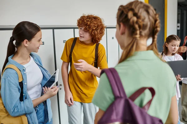Підліткові однокласники спілкуються в шкільному коридорі, студенти-підлітки тримають пристрої, молодіжну культуру, навчаються — стокове фото