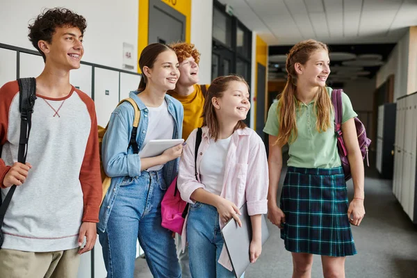 Счастливые школьники-подростки, смотрящие в сторону и стоящие вместе в школьном коридоре, одноклассники — стоковое фото