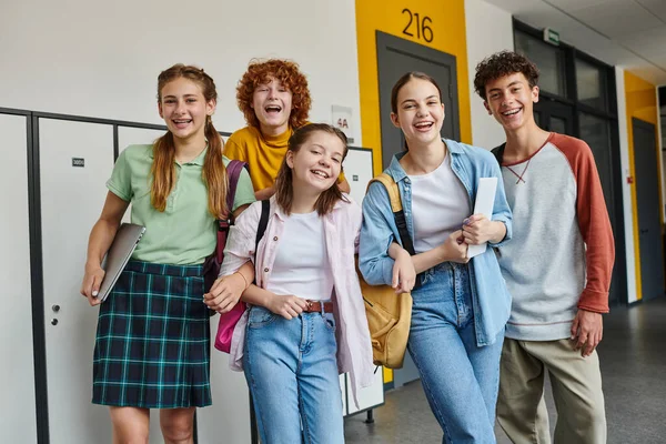 Teenage schoolkids looking at camera and standing in school hallway, teen classmates with devices — Stock Photo