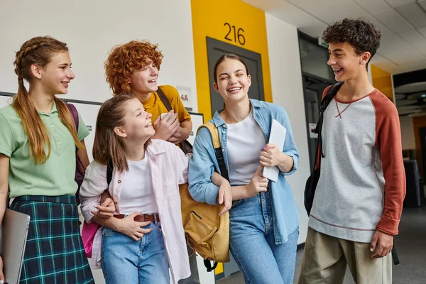 Школьники-подростки смеются и стоят вместе в школьном коридоре, одноклассники-подростки с устройствами — стоковое фото