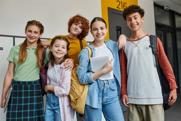 De vuelta a la escuela, adolescentes felices mirando la cámara en el pasillo, sosteniendo los dispositivos y sonriendo juntos - foto de stock
