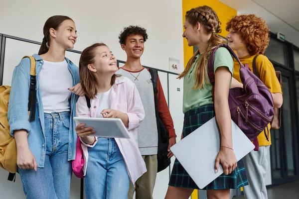 Девочка-подросток указывает на цифровой планшет и смотрит на одноклассников, подростков в школьном коридоре — стоковое фото