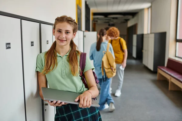 Позитивна дівчина-підліток тримає ноутбук і дивиться на камеру в коридорі, назад до концепції школи — стокове фото