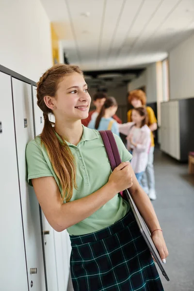 Cheerful teenage girl holding laptop and looking away in hallway, back to school concept, modern age — Stock Photo