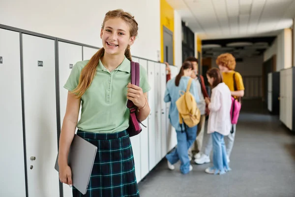 Cheerful teenage girl holding laptop and looking away in hallway, blur, schoolkids on background — Stock Photo