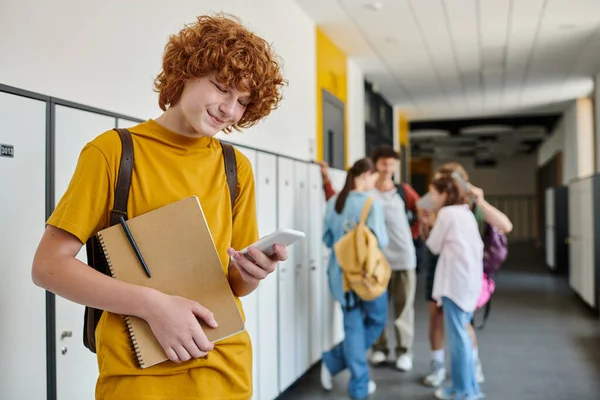 Happy redhead schoolboy using smartphone and holding notebook, blur, students in school hallway — Stock Photo