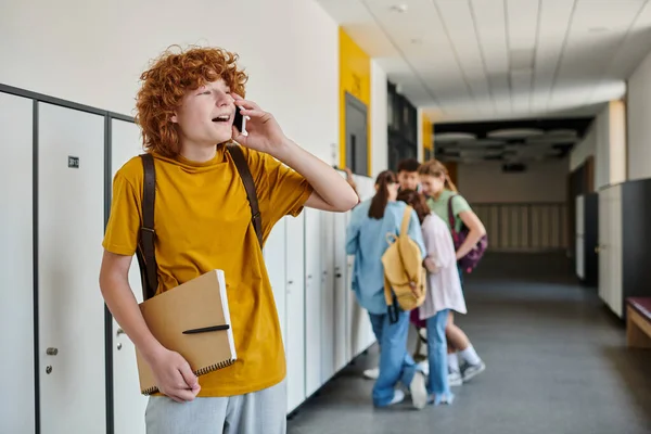 Llamada telefónica, colegial pelirroja hablando en el teléfono inteligente, estudiante feliz en el pasillo de la escuela durante el descanso - foto de stock