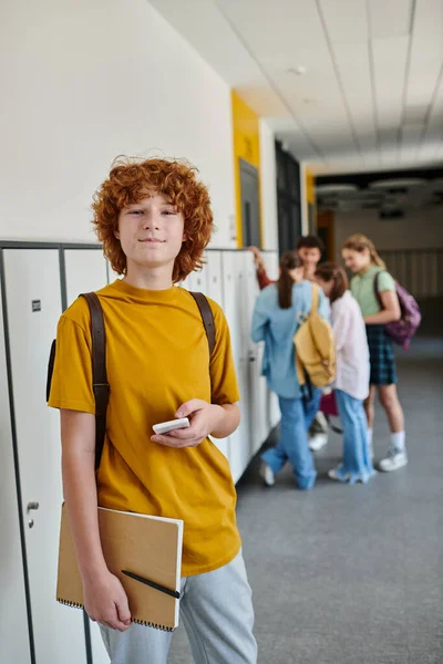 Pelirroja escolar sosteniendo teléfono inteligente, estudiante feliz en el pasillo de la escuela mirando a la cámara durante el descanso - foto de stock