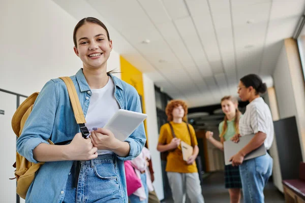 Teenage girl with digital tablet looking at camera in school hallway, blurred background, students — Stock Photo