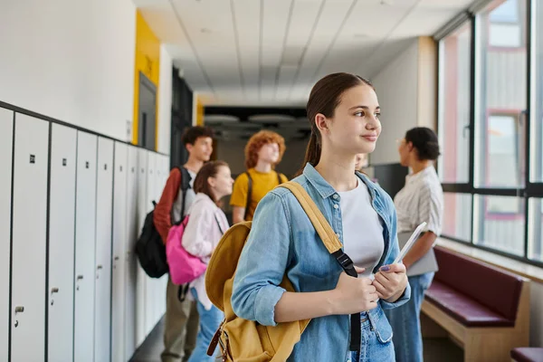 Adolescente con tableta digital mirando hacia otro lado en el pasillo de la escuela, borrosa, estudiantes y maestro - foto de stock