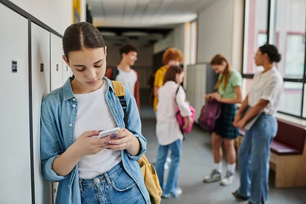 Дівчина-підліток смс на смартфоні в шкільному коридорі, студенти і вчителька на розмитому фоні — стокове фото