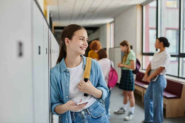 Feliz adolescente sosteniendo teléfono inteligente en el pasillo de la escuela, diversidad cultural, maestro y niños, desenfoque - foto de stock