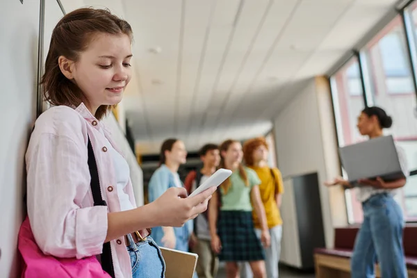 Fille heureuse utilisant un smartphone, bavarder et debout dans le couloir de l'école, diversité, enseignant, enfants, flou — Photo de stock
