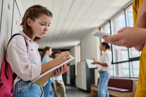 Studentessa concentrata prendendo appunti, tenendo il taccuino vicino al ragazzo utilizzando lo smartphone nel corridoio della scuola — Foto stock