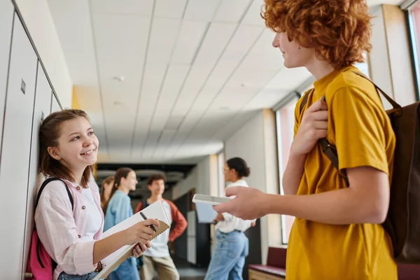 Heureuse écolière tenant carnet et regardant garçon avec smartphone, couloir de l'école, pause, flou — Photo de stock