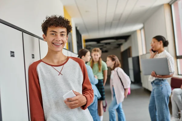 Ragazzo felice con l'apparecchio acustico che tiene smartphone e guarda la fotocamera durante la pausa nel corridoio della scuola — Foto stock