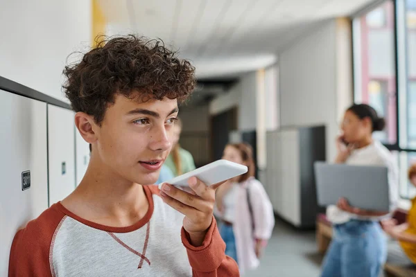 Adolescente escolar grabación de mensaje de voz, celebración de teléfono inteligente durante el descanso en el pasillo de la escuela, desenfoque - foto de stock