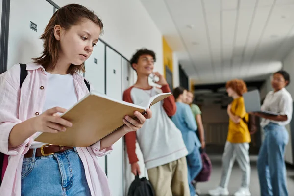 Schoolgirl holding notebook and standing near blurred and happy teen classmate, students, hallway — Stock Photo