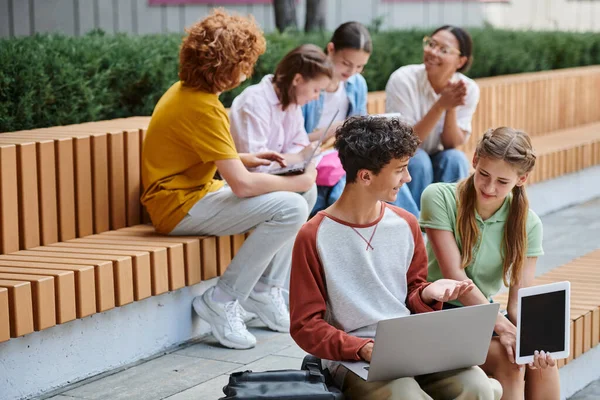 De vuelta a la escuela, niño adolescente feliz mostrando portátil a su compañero de clase, estudiantes y profesores al aire libre, diversidad - foto de stock