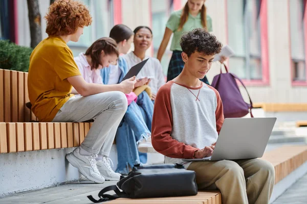 Повернутися до школи, розумний хлопчик-підліток використовує ноутбук поруч з однокласниками на відкритому повітрі, різноманітність, студенти — стокове фото