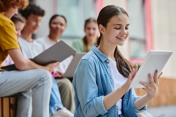 Счастливая девочка-подросток улыбается и с помощью цифрового планшета рядом размытые одноклассники и учитель, разнообразие — стоковое фото