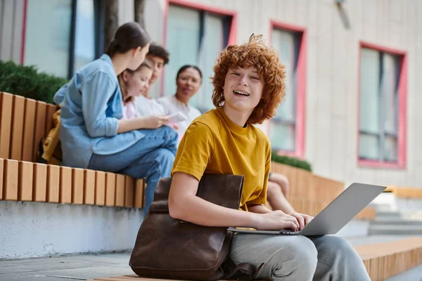 De vuelta a la escuela, chico pelirrojo feliz con el pelo rizado usando el ordenador portátil cerca de compañeros de clase y maestro, borroso - foto de stock