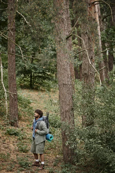 Jeune homme indien avec sac à dos, bâtons de trekking et tapis de fitness debout entre les arbres dans la forêt — Photo de stock