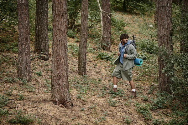 Sonriente joven senderista indio con mochila mirando hacia otro lado mientras camina entre árboles en el bosque - foto de stock
