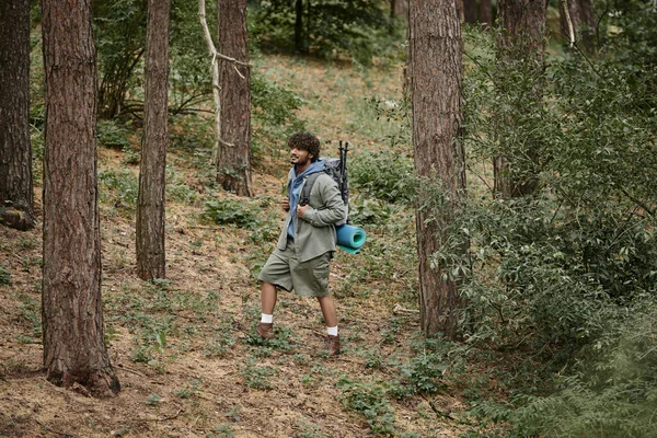 Giovane escursionista indiano in camicia e pantaloncini che tiene lo zaino mentre cammina nella foresta — Foto stock