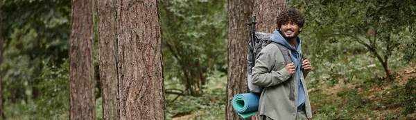 Веселий молодий індійський пішохід з рюкзаком, що стоїть між деревами в лісі, банер — стокове фото