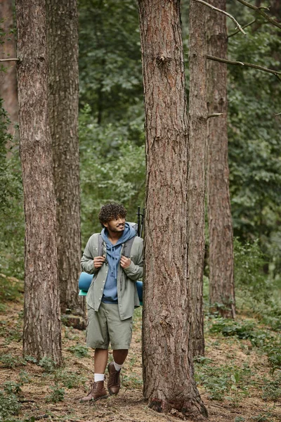 Улыбающийся молодой индийский путешественник с рюкзаком, стоящим рядом с деревом во время отдыха в лесу — стоковое фото
