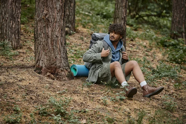 Молодий кучерявий індійський пішохід з рюкзаком, що сидить на землі біля дерев у лісі — стокове фото