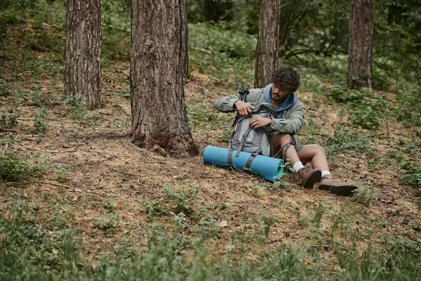Curly young indian traveler opening backpack while resting and sitting on ground in forest — Stock Photo