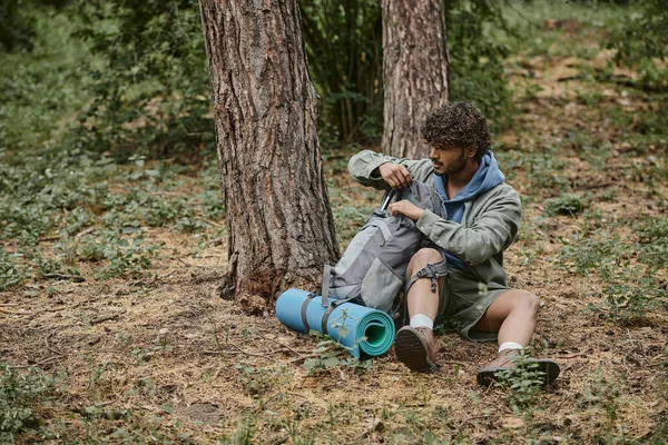 Young indian hiker opening backpack near fitness mat while resting on ground in forest — Stock Photo