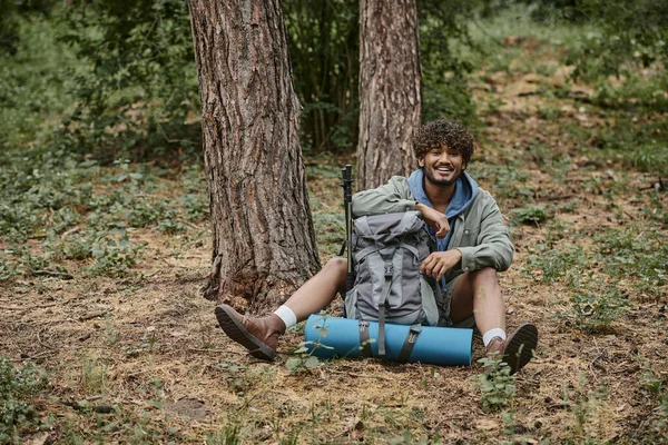 Cheerful young indian man looking at camera near backpack on ground in forest — Stock Photo