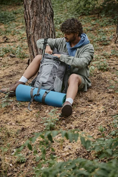 Joven viajero indio abriendo mochila con bastones de trekking y colchoneta de fitness en el bosque - foto de stock