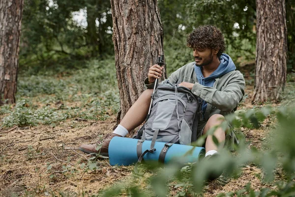 Positive young indian hiker taking trekking poles from backpack on ground in forest — Stock Photo