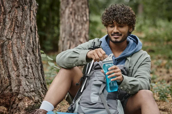 Sonriente joven senderista indio sosteniendo botella deportiva cerca de la mochila mientras descansa en el bosque - foto de stock