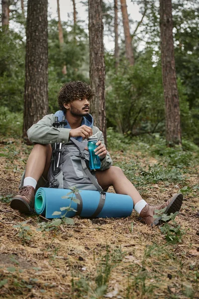 Junger indischer Reisender mit Sportflasche in der Nähe von Rucksack mit Wanderstöcken im Wald — Stockfoto