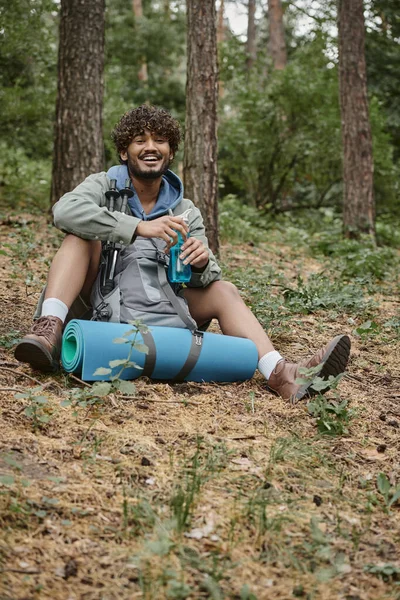 Senderista indio despreocupado sosteniendo botella deportiva cerca de la mochila y bastones de trekking en el suelo en el bosque - foto de stock