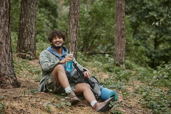Alegre joven turista indio sosteniendo botella deportiva cerca de la mochila en el suelo en el bosque - foto de stock