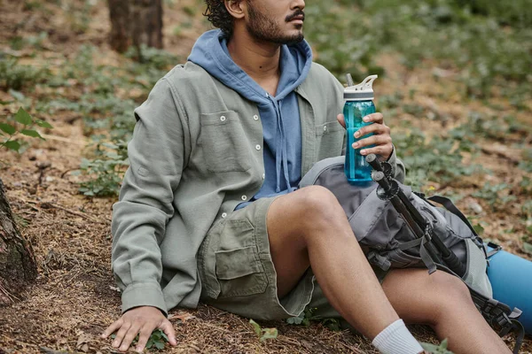 Abgeschnittene Ansicht eines jungen indischen Mannes mit Sportflasche, während er in der Nähe von Rucksack im Wald sitzt — Stockfoto
