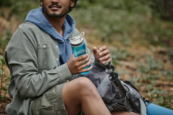 Vista cortada de viajante indiano segurando garrafa de esportes perto de mochila na floresta turva — Fotografia de Stock