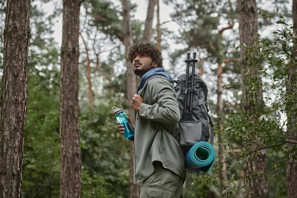 Young curly indian tourist holding backpack and sports bottle in forest — Stock Photo